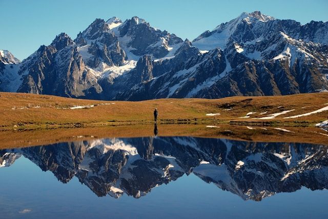 A person stands on a grassy shore before a still lake, which reflects a large, snow-covered mountain range under a clear sky.