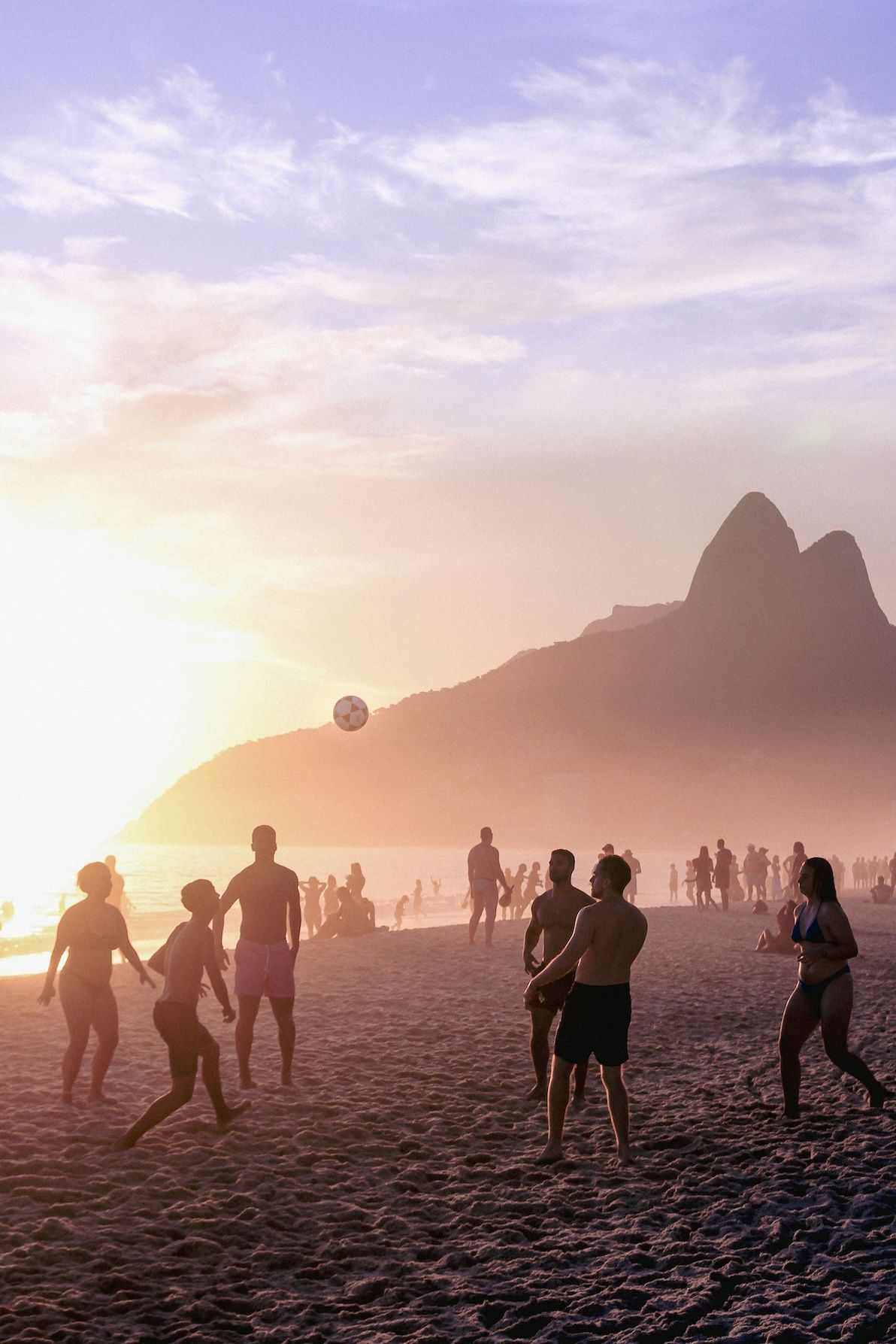 Sagome di un gruppo di persone che giocano a palla su una spiaggia sabbiosa al tramonto, con montagne lontane oltre l'acqua.