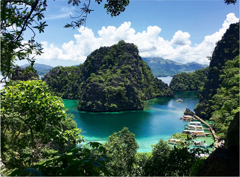A high-angle view of a turquoise lagoon surrounded by steep, jungle-covered islands, with boats moored at a wooden dock.