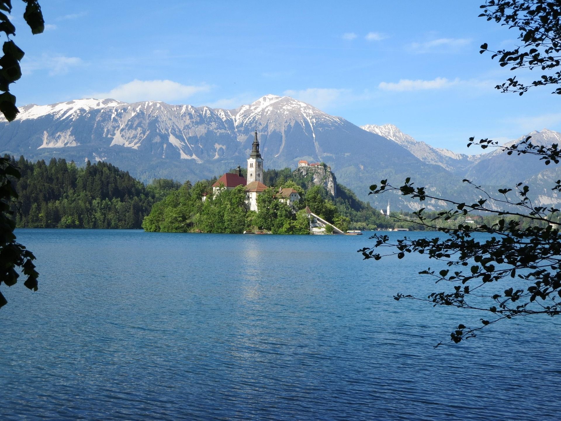 Una chiesa su una piccola isola alberata in un lago, con montagne innevate sullo sfondo sotto un cielo blu.