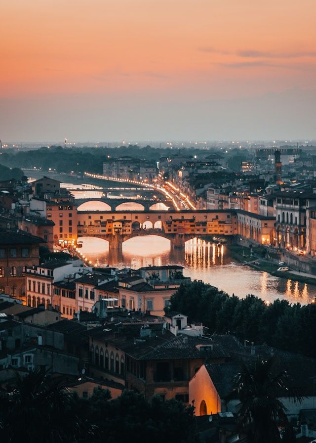 An illuminated stone bridge crosses a river in a dense cityscape at dusk, its lights reflecting on the water.