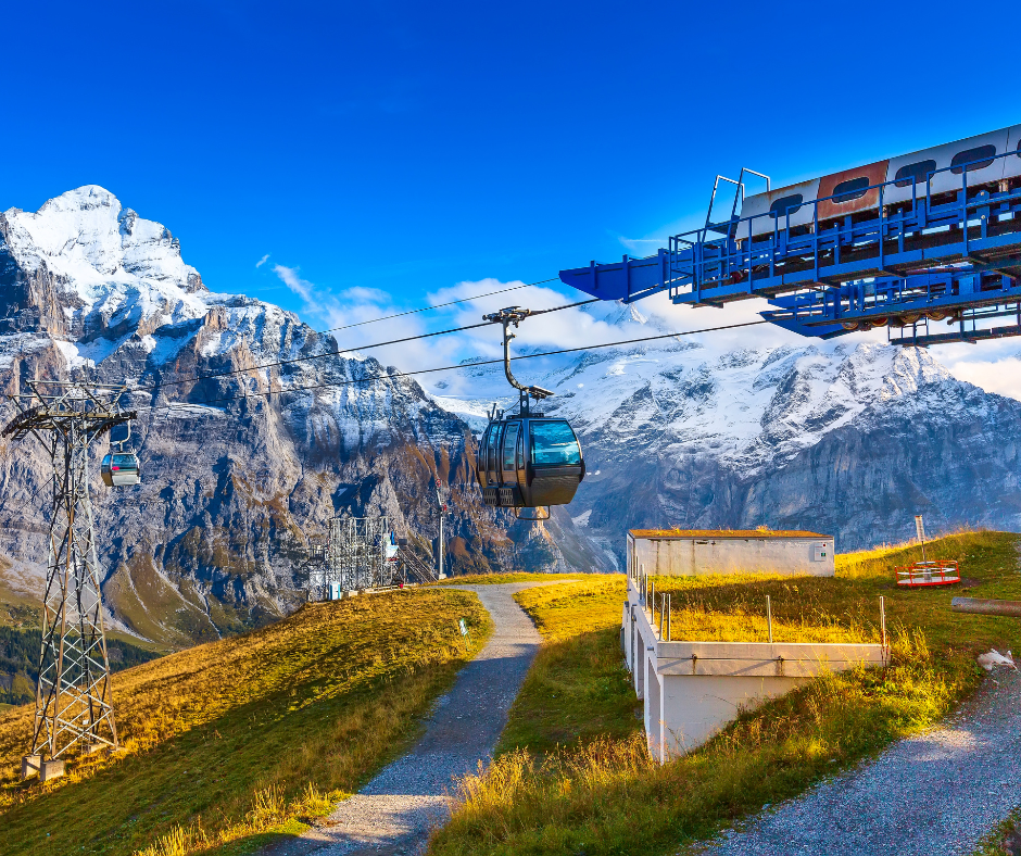 A cable car travels over a green mountain slope, with towering snow-capped peaks visible against a clear blue sky.