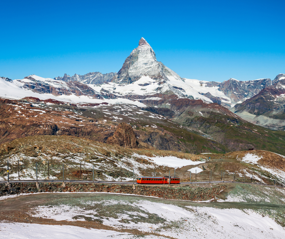 A red train travels on a track across a rocky, partially snow-covered mountainside, with a prominent snow-capped peak in the background.