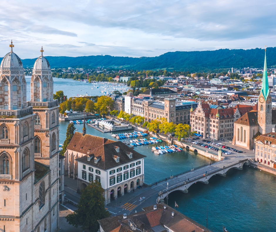 An aerial view of a historic city with church spires and a bridge over a river, with a large lake and hills in the background.