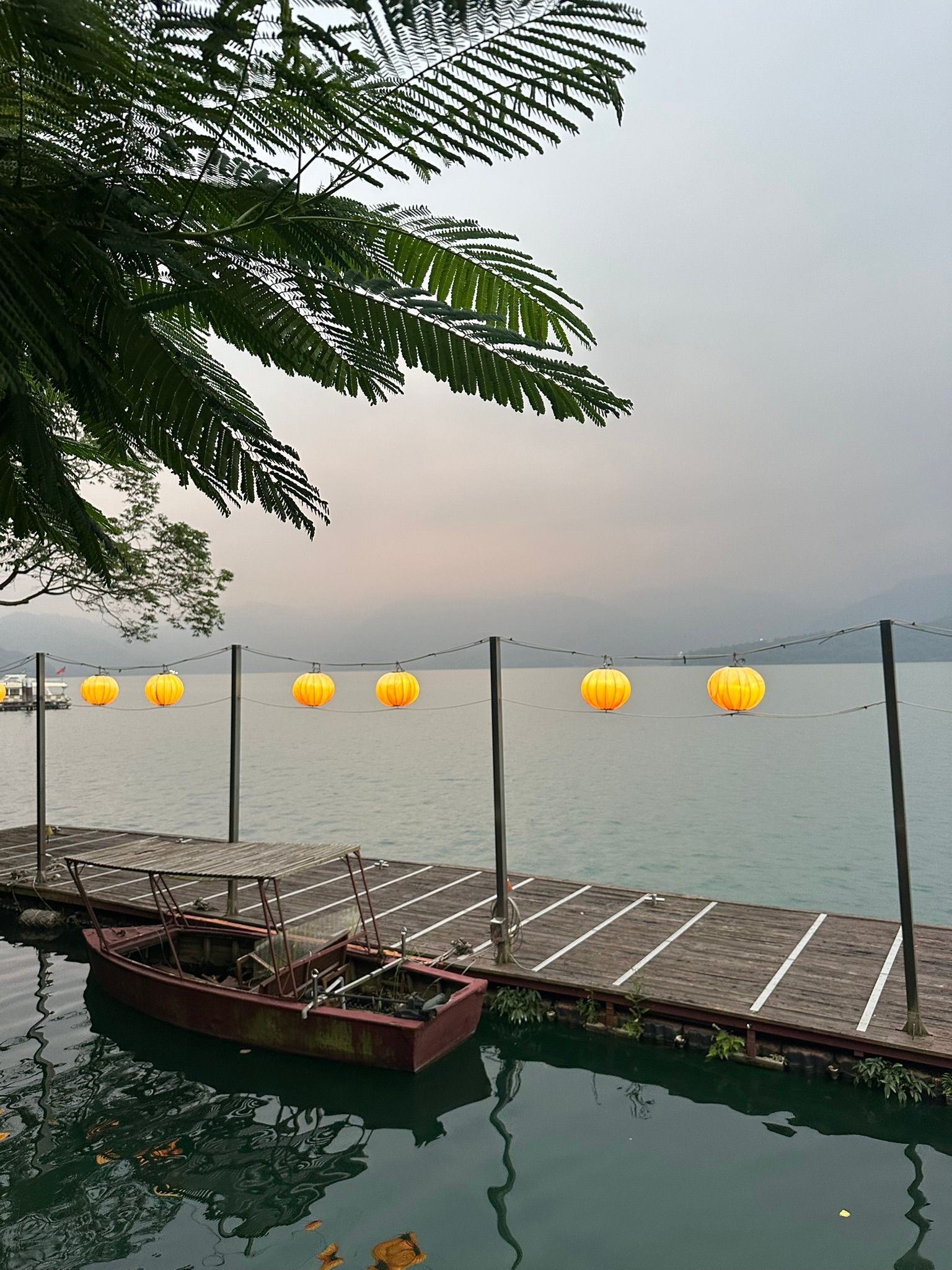 Leuchtende gelbe Papierlaternen hängen über einem Holzsteg, an dem ein kleines Boot auf einem ruhigen See vertäut ist.