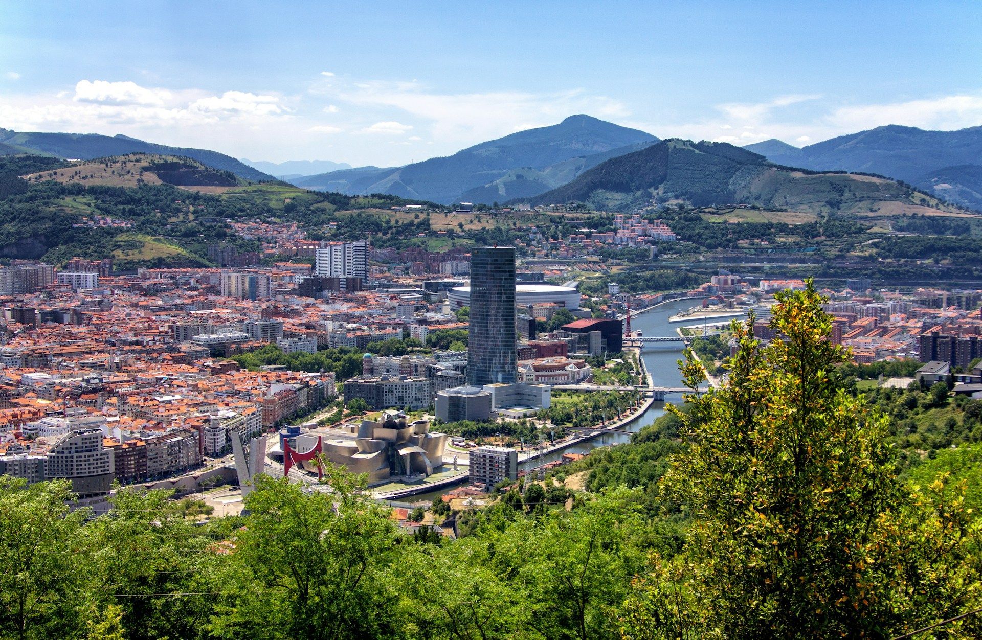 Una veduta panoramica di una grande città con un fiume serpeggiante, incastonata tra dolci colline verdi e montagne sotto un cielo blu.