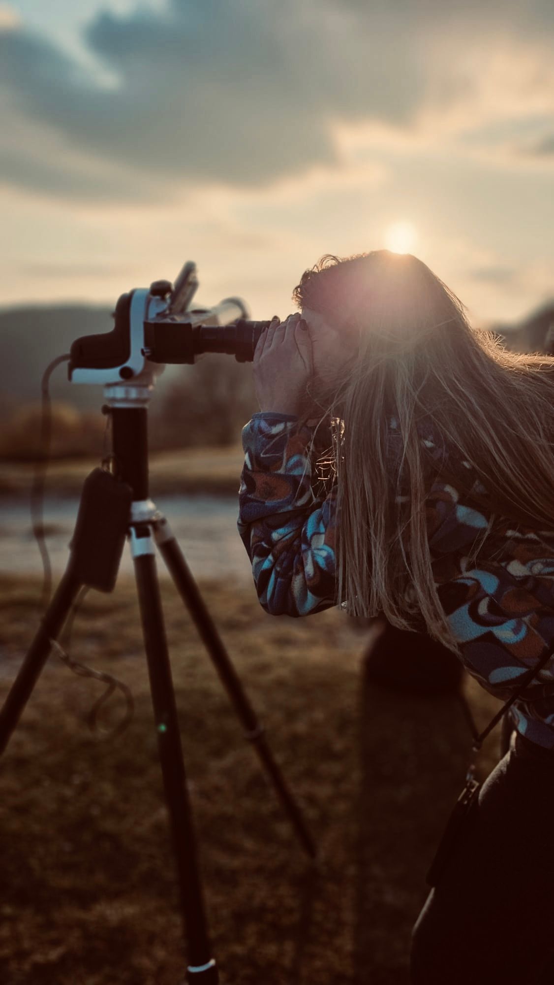 Una persona con i capelli lunghi guarda attraverso una fotocamera montata su un treppiede, all'aperto al tramonto, con il sole che crea un bagliore.
