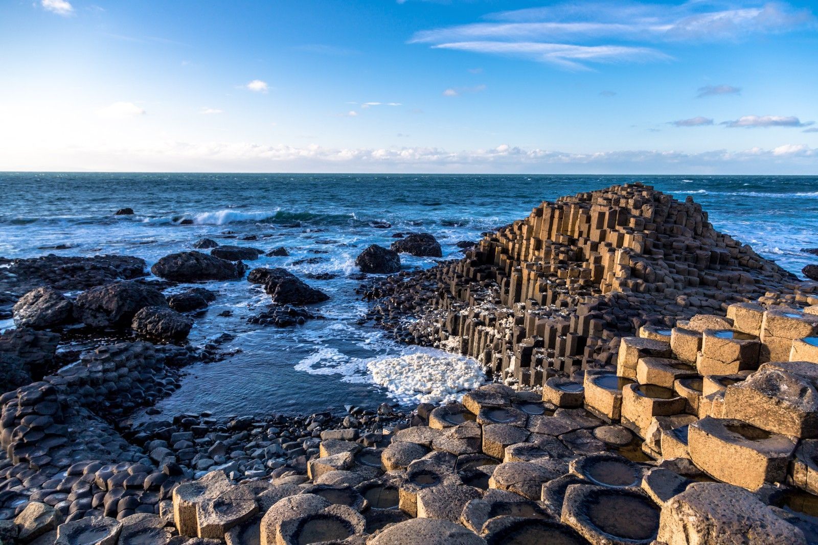 Colonne esagonali di basalto incastrate formano un paesaggio costiero, con le onde dell'oceano che lambiscono la riva sotto un cielo azzurro.