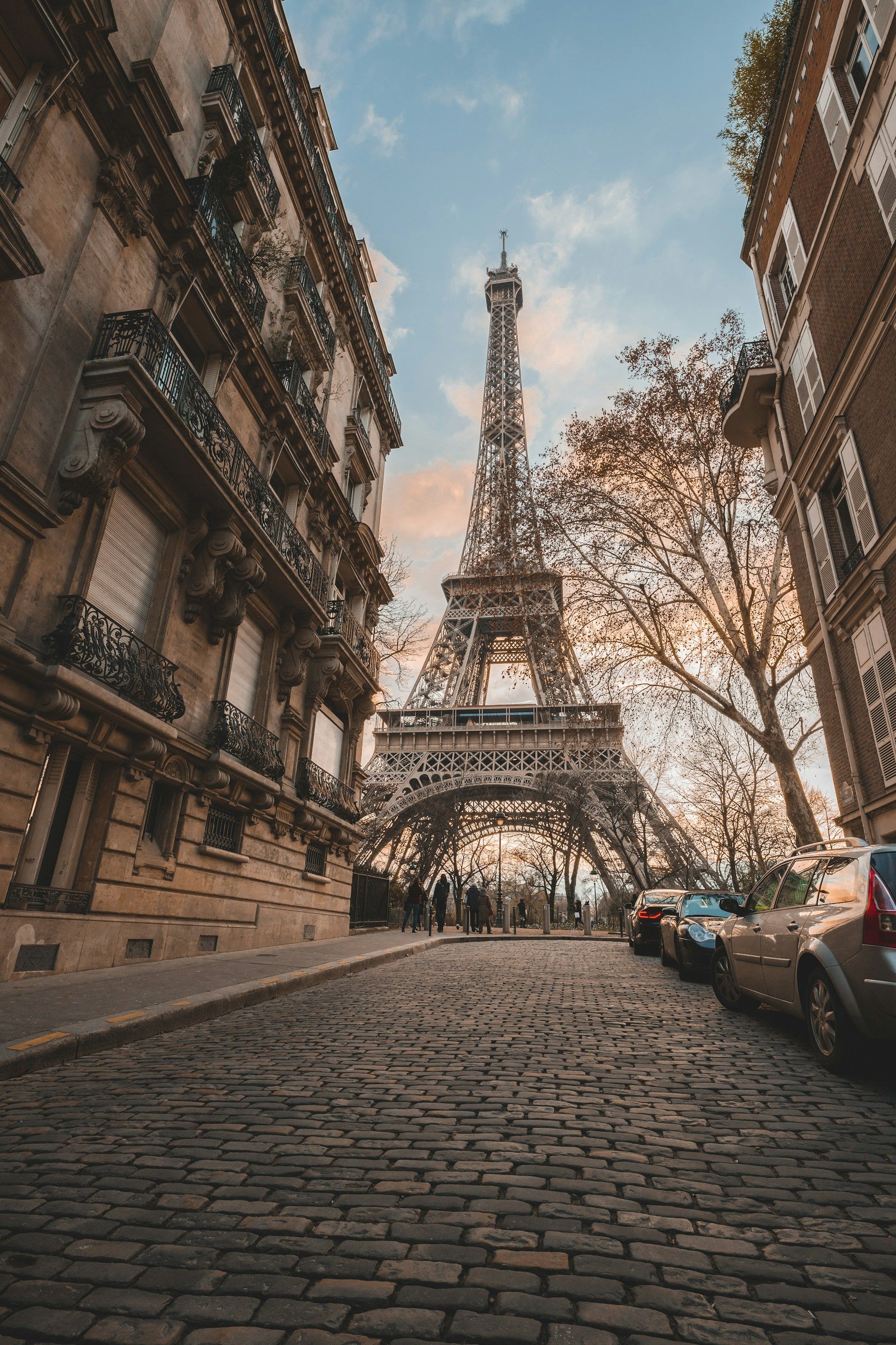 Una vista dal basso della Torre Eiffel da una strada acciottolata, incorniciata da edifici tradizionali e alberi sotto un cielo nuvoloso.