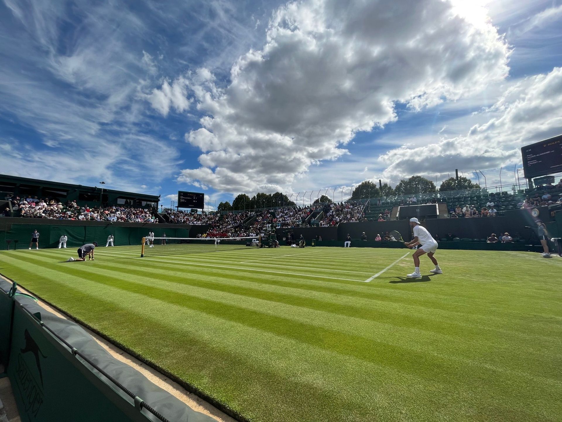 Inquadratura dal basso di una partita di tennis su campo in erba a strisce, con un giocatore in bianco pronto a giocare sotto un cielo azzurro nuvoloso.