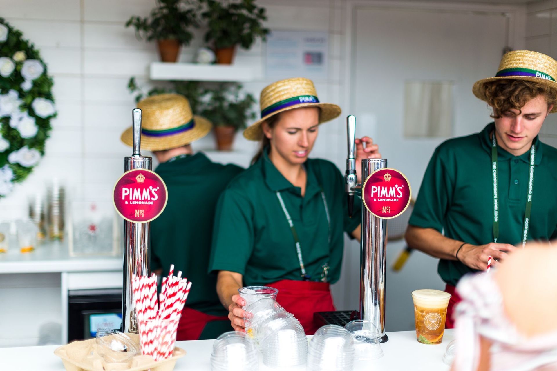Bartender in uniformi verdi e cappelli di paglia versano bevande dai rubinetti di Pimm's & Lemonade a una bancarella.