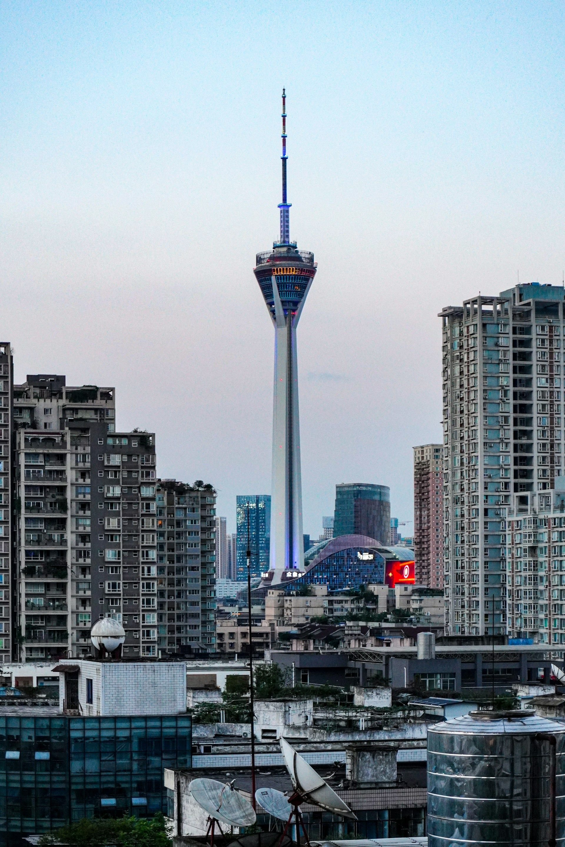 Una torre di osservazione alta e moderna si erge sopra una fitta città, piena di edifici residenziali, sotto un cielo pallido.