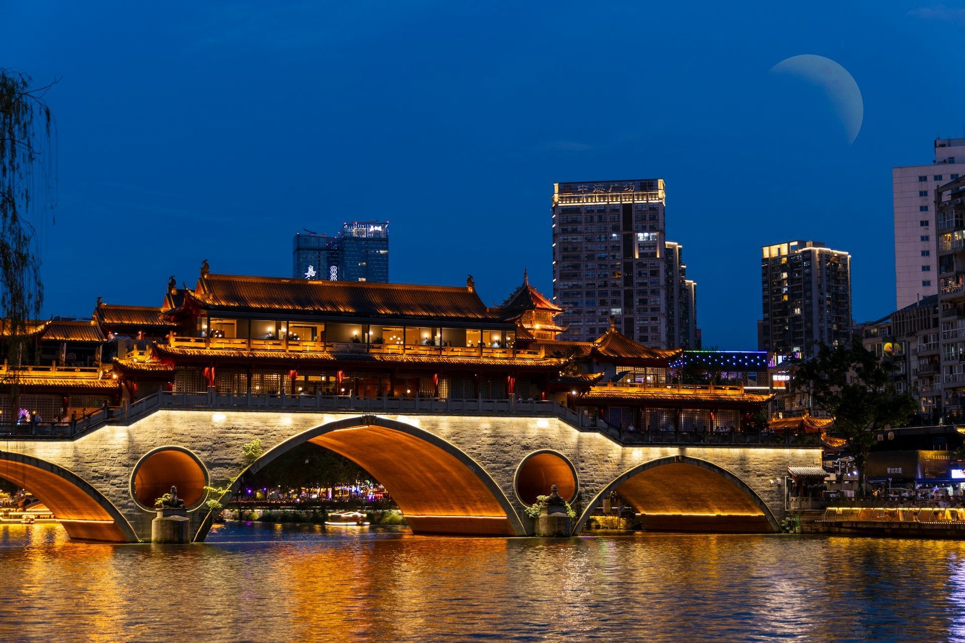 Un ponte tradizionale cinese illuminato con edifici in cima attraversa un fiume di notte, con moderni grattacieli sullo sfondo sotto una luna crescente.