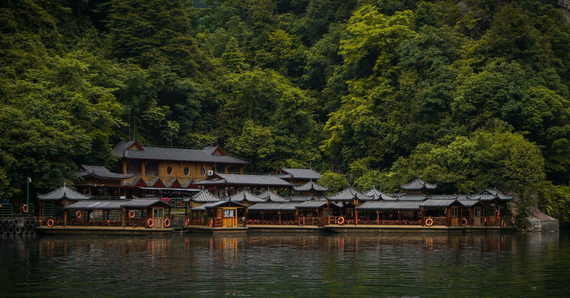 Barche turistiche con tetto a pagoda sono ormeggiate su un fiume tranquillo, davanti a una collina verde e lussureggiante, coperta di boschi.