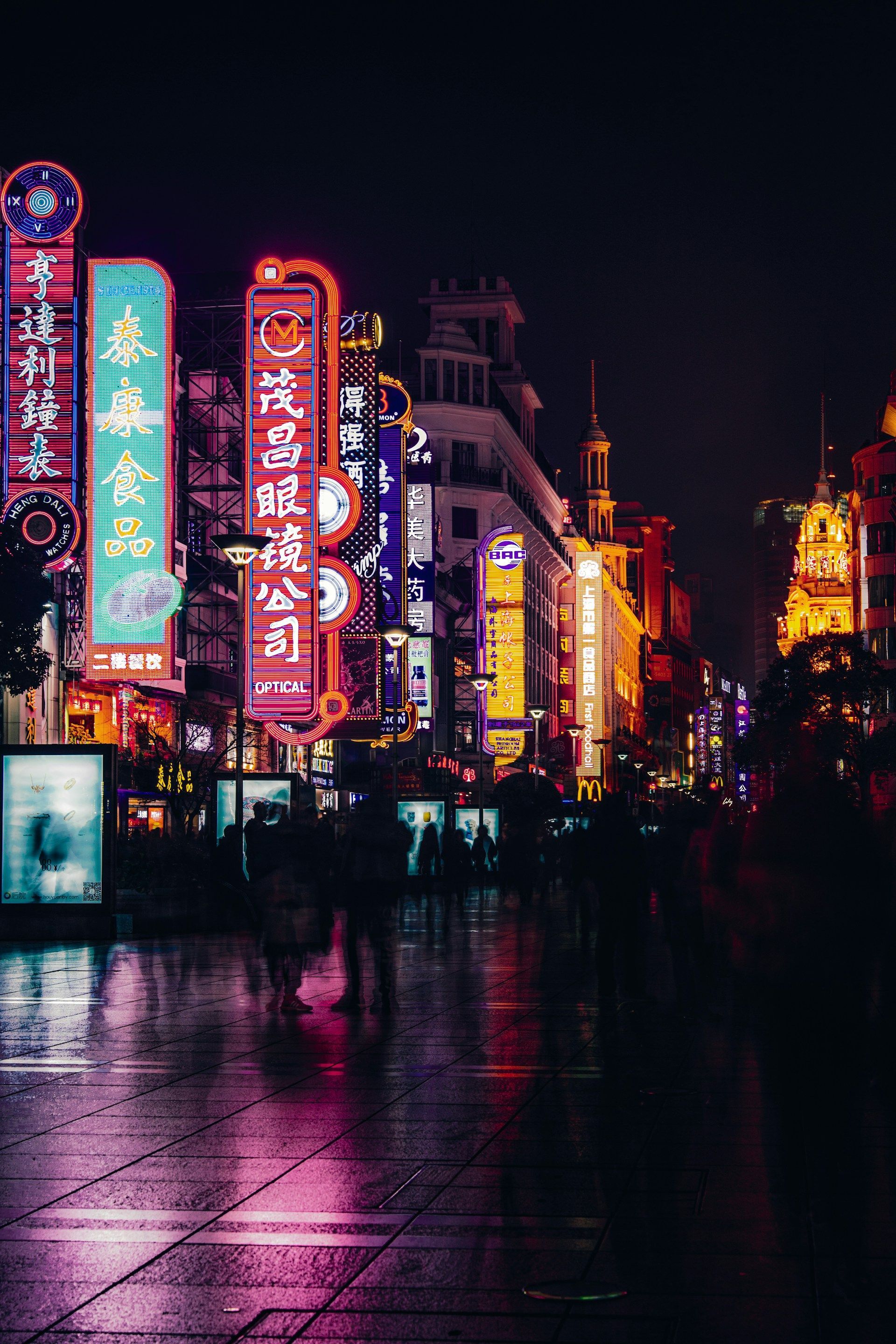 Di notte, una strada cittadina bagnata riflette il bagliore di vivaci insegne al neon verticali con caratteri asiatici, mentre la gente passeggia.