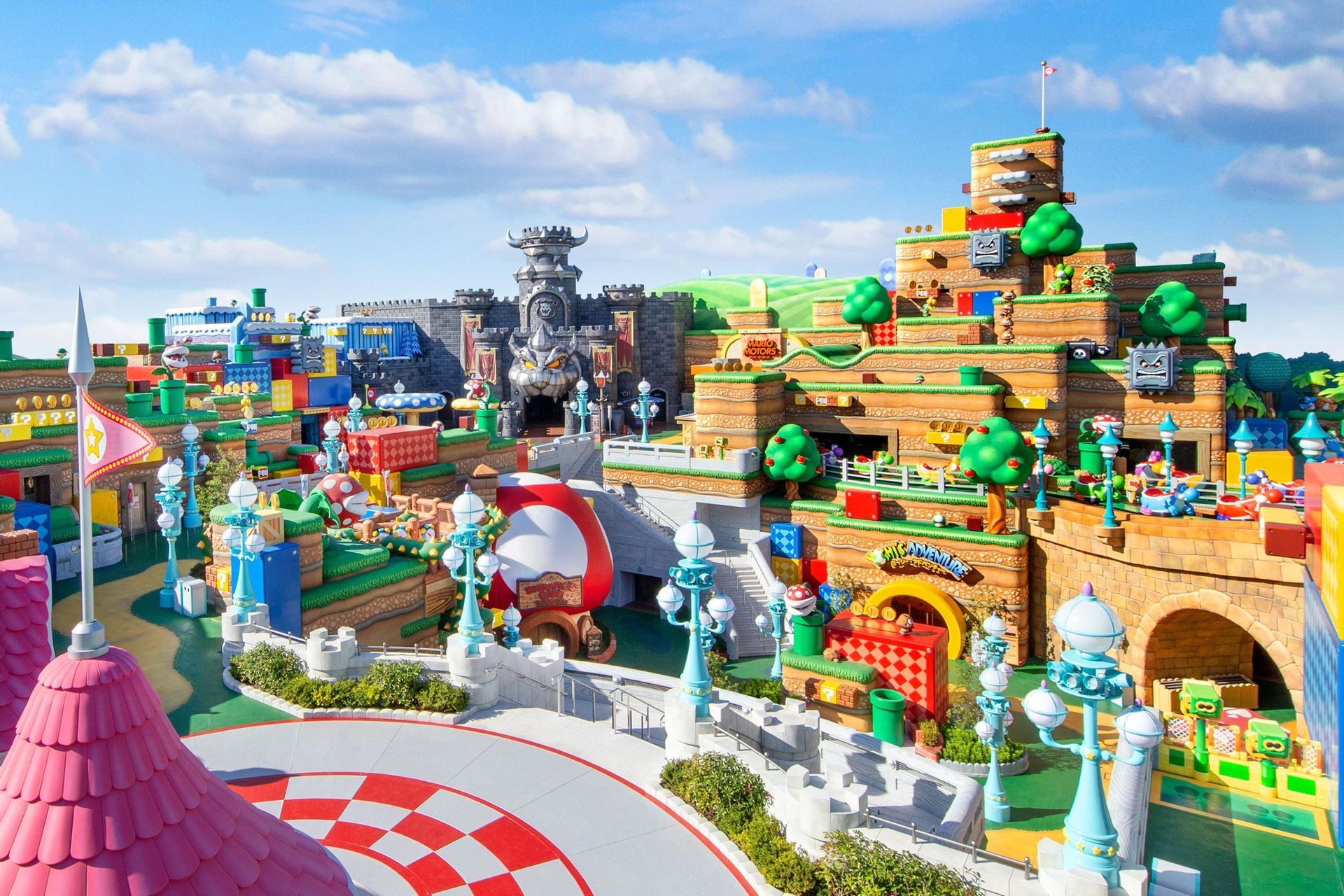 Una vista panoramica su un colorato parco divertimenti, progettato per sembrare un livello del videogioco di Super Mario, sotto un cielo azzurro parzialmente nuvoloso.