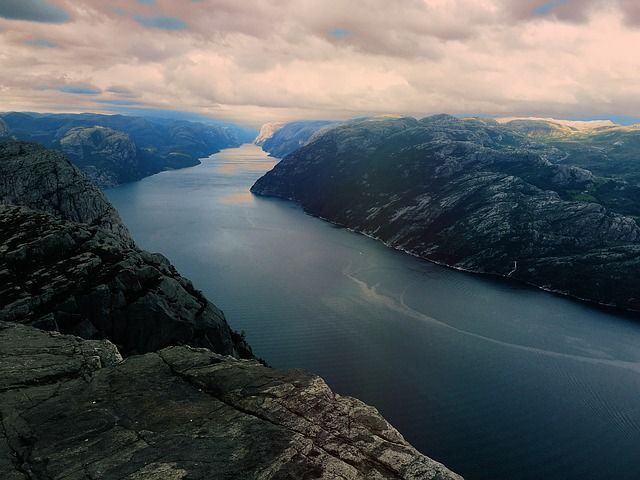 Una vista dall'alto da una scogliera rocciosa che domina un vasto fiordo che si snoda tra ripide montagne sotto un cielo nuvoloso.