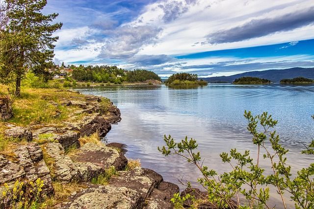 Una costa rocciosa incontra un lago calmo con diverse piccole isole alberate e colline lontane sotto un cielo parzialmente nuvoloso.