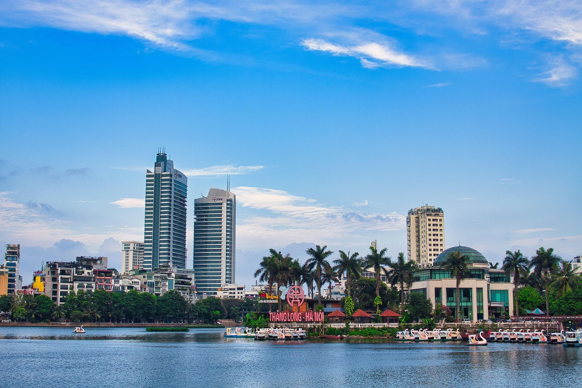 Uno skyline cittadino con alti edifici e alberi verdi, visto attraverso uno specchio d'acqua con pedalò a forma di cigno ormeggiati lungo la riva.