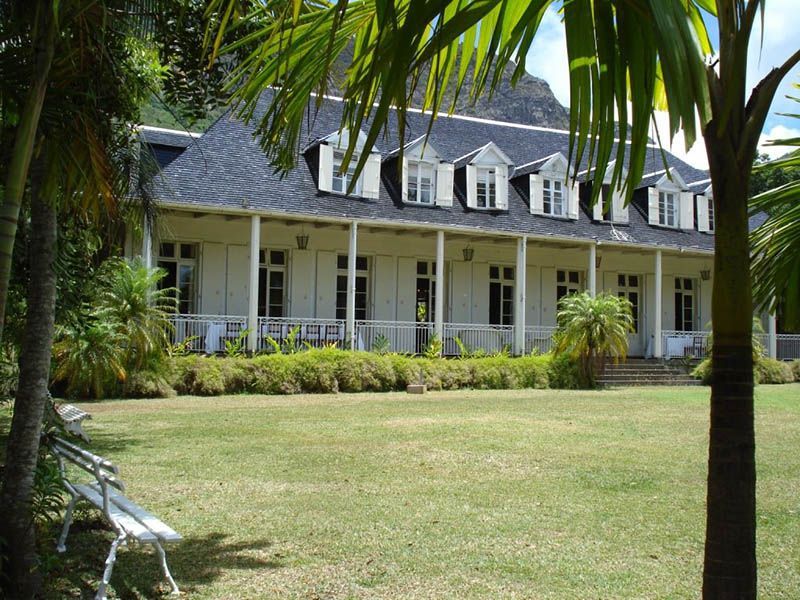 Una casa bianca in stile coloniale con tetto scuro e veranda si erge dietro un ampio prato verde, incorniciata da lussureggianti palme.