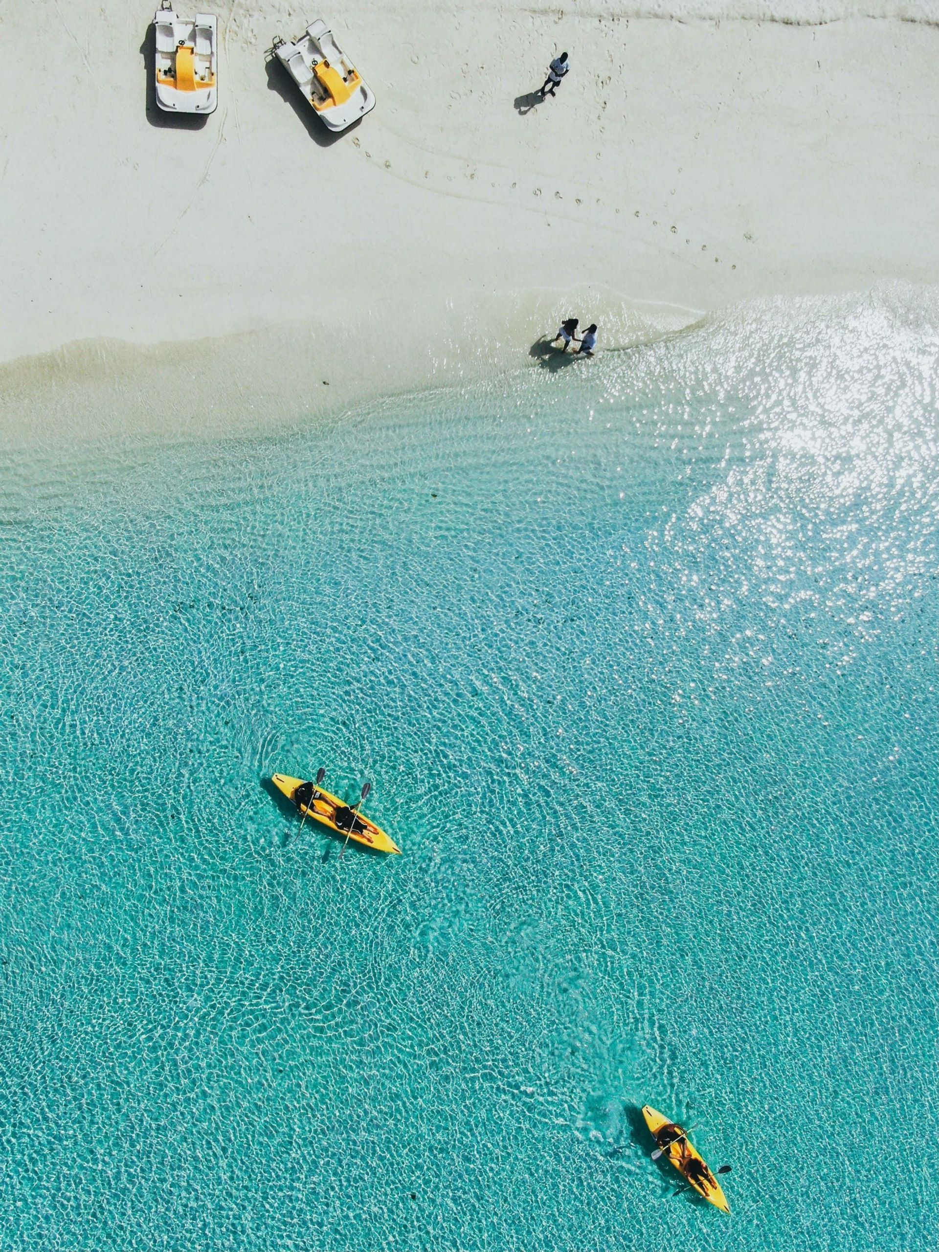 Vista aerea di due kayak gialli in acque limpide e turchesi accanto a una spiaggia di sabbia bianca con persone e pedalò.