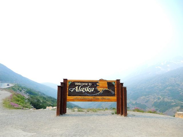 A wooden 'Welcome to Alaska' sign stands on a gravel patch overlooking a winding mountain road and hazy peaks.