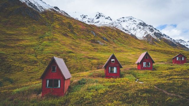 Four small red A-frame cabins are scattered on a grassy hillside at the base of a large, snow-capped mountain under a cloudy sky.