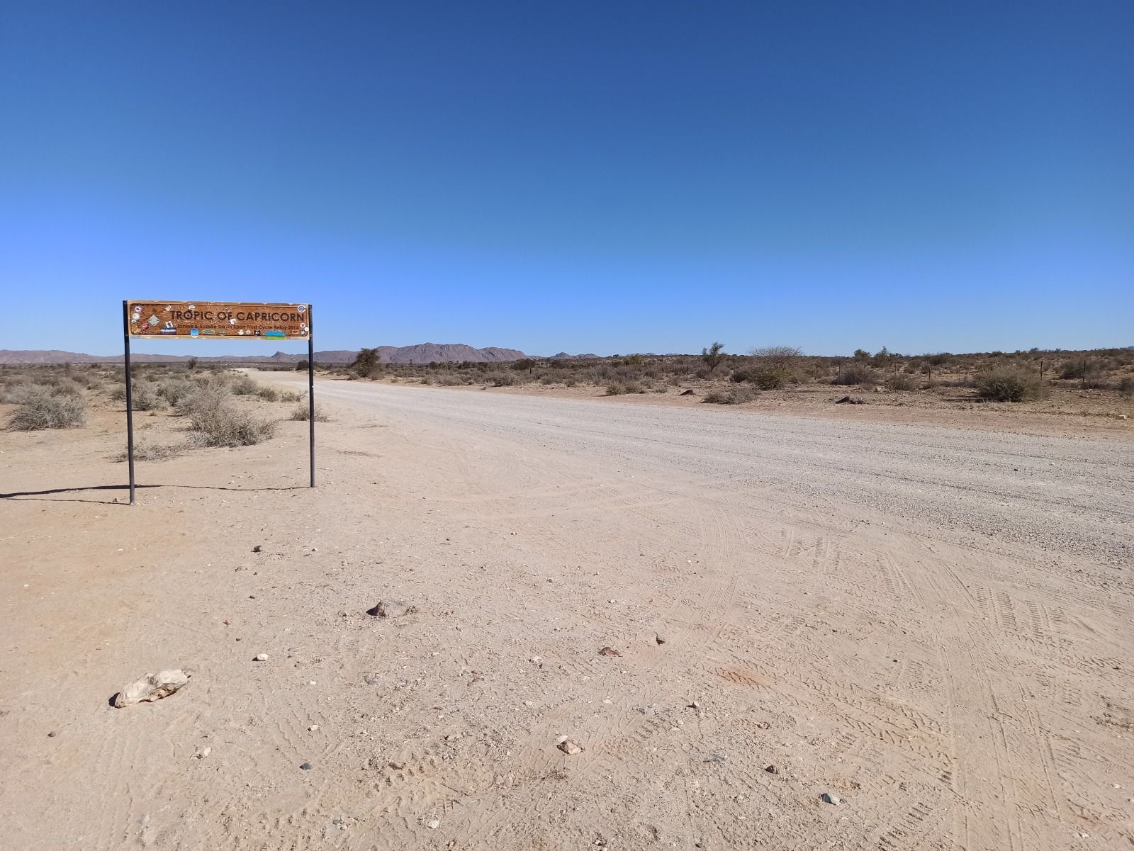 Un cartello 'Tropico del Capricorno' si erge accanto a una lunga strada sterrata in un vasto paesaggio desertico sotto un cielo azzurro.