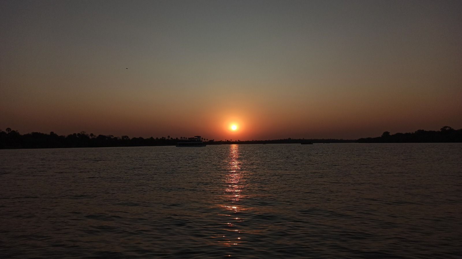 Il sole tramonta su un ampio specchio d'acqua, la sua luce arancione si riflette sulla superficie, con una barca visibile in lontananza.