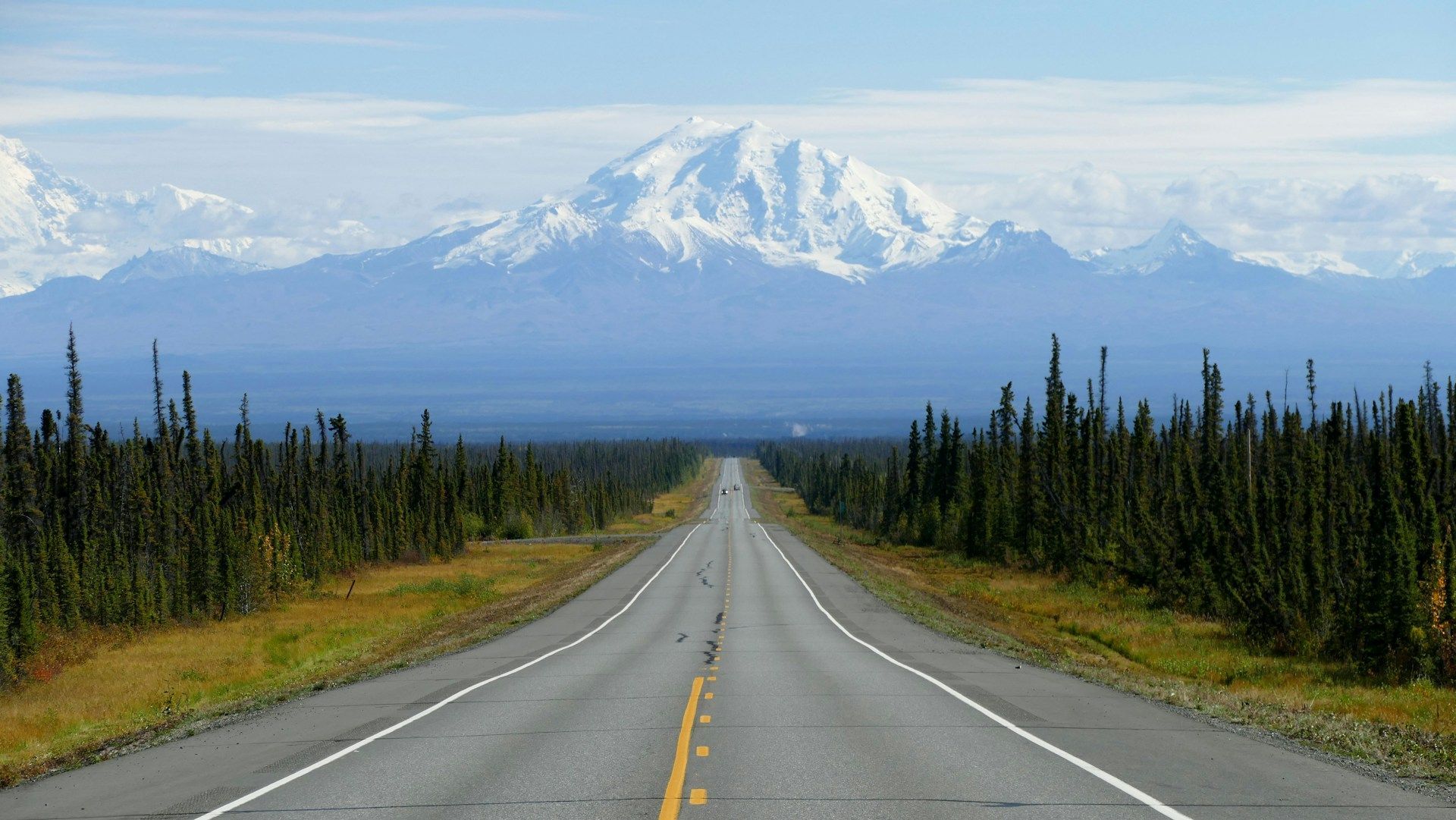 A straight paved road stretches through a dense evergreen forest towards a large, snow-covered mountain range under a partly cloudy sky.