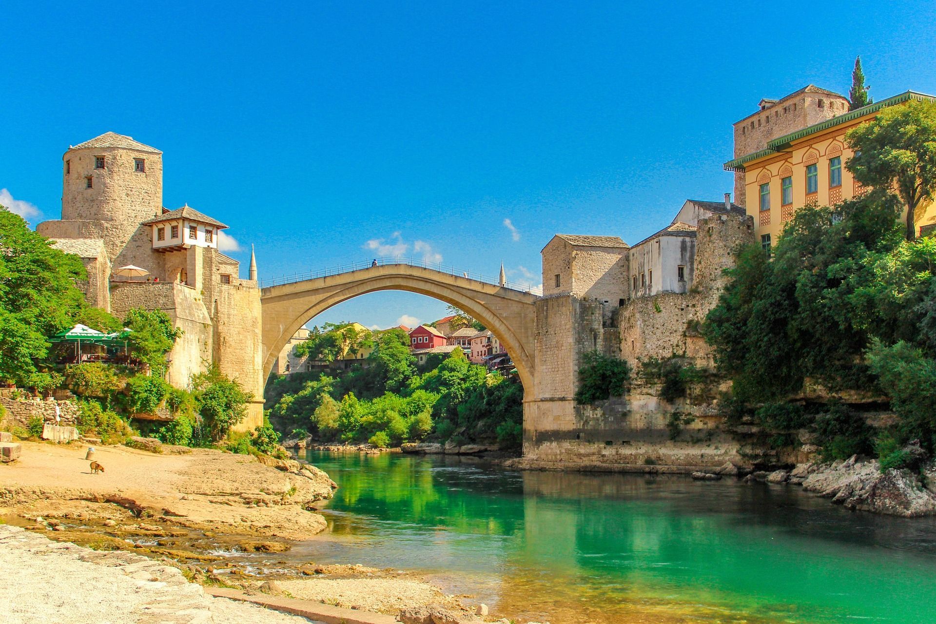 Un ponte ad arco in pietra attraversa un fiume turchese, fiancheggiato da edifici storici e una vegetazione lussureggiante, sullo sfondo di un cielo azzurro luminoso.