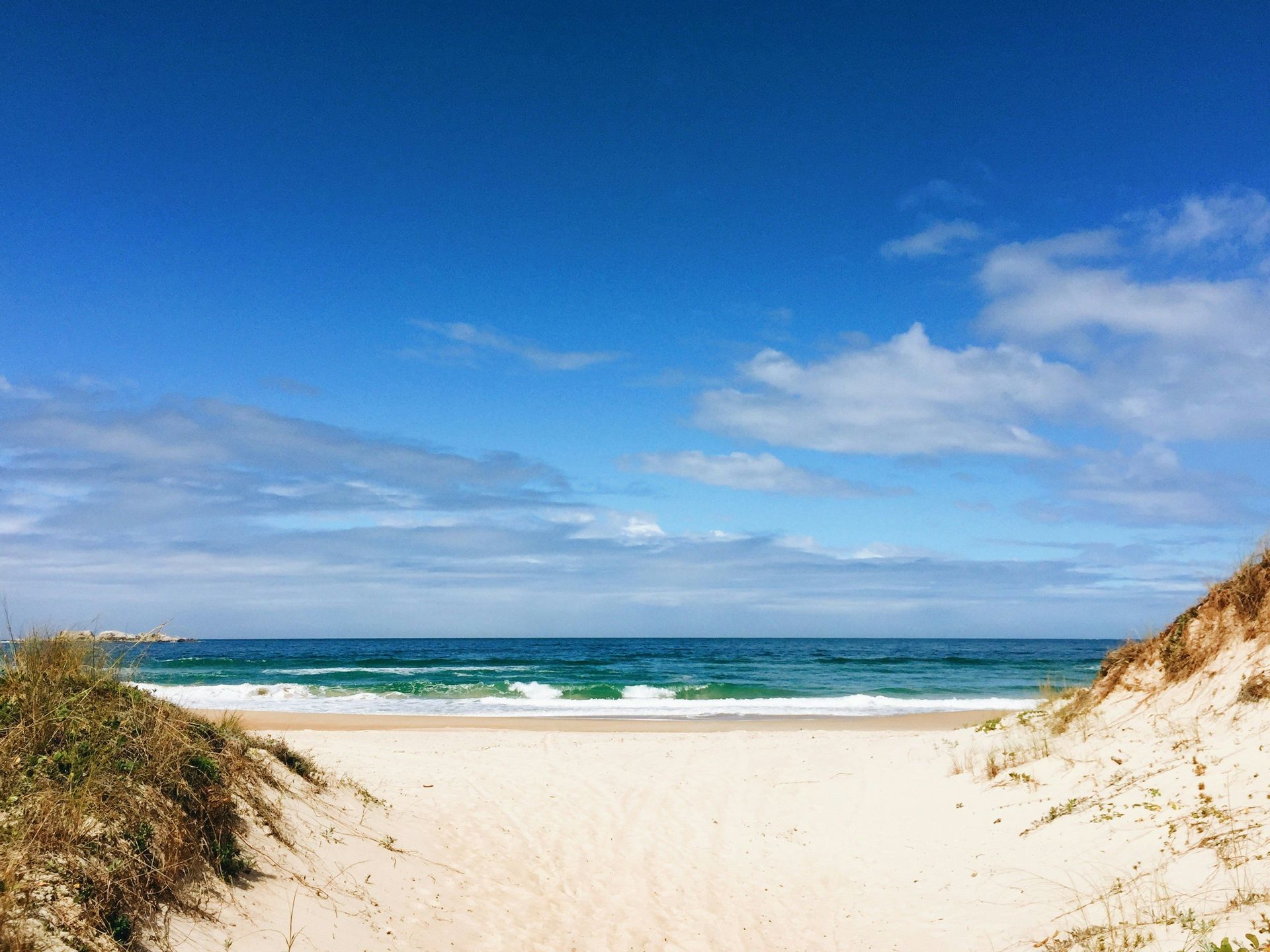 Un sentiero sabbioso tra due dune erbose conduce a una spiaggia di sabbia bianca con onde blu dell'oceano sotto un cielo parzialmente nuvoloso.
