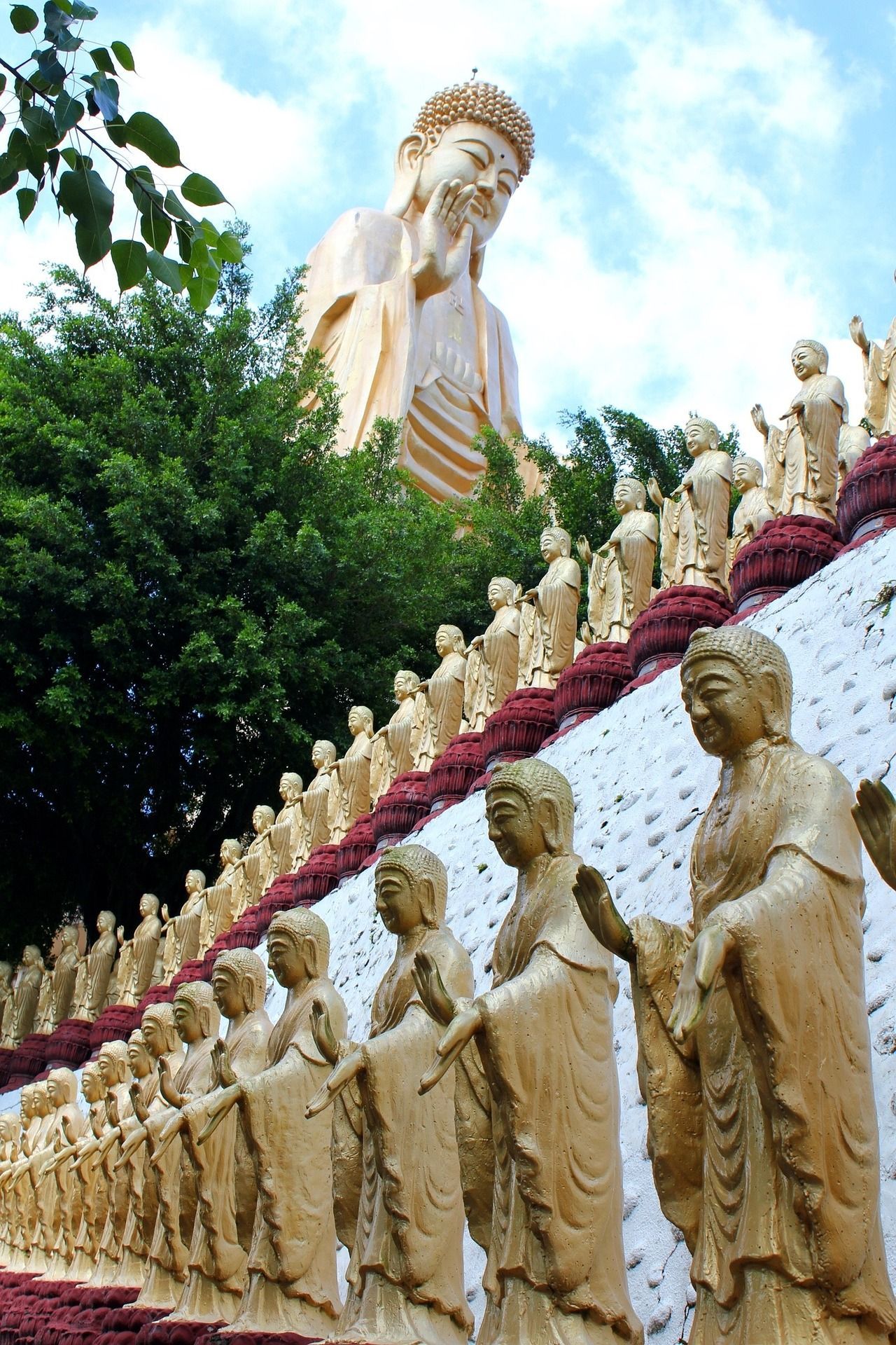 Eine riesige goldene Buddha-Statue thront über vielen kleineren goldenen Statuen, die in Reihen an einem Berghang unter einem teilweise bewölkten Himmel angeordnet sind.