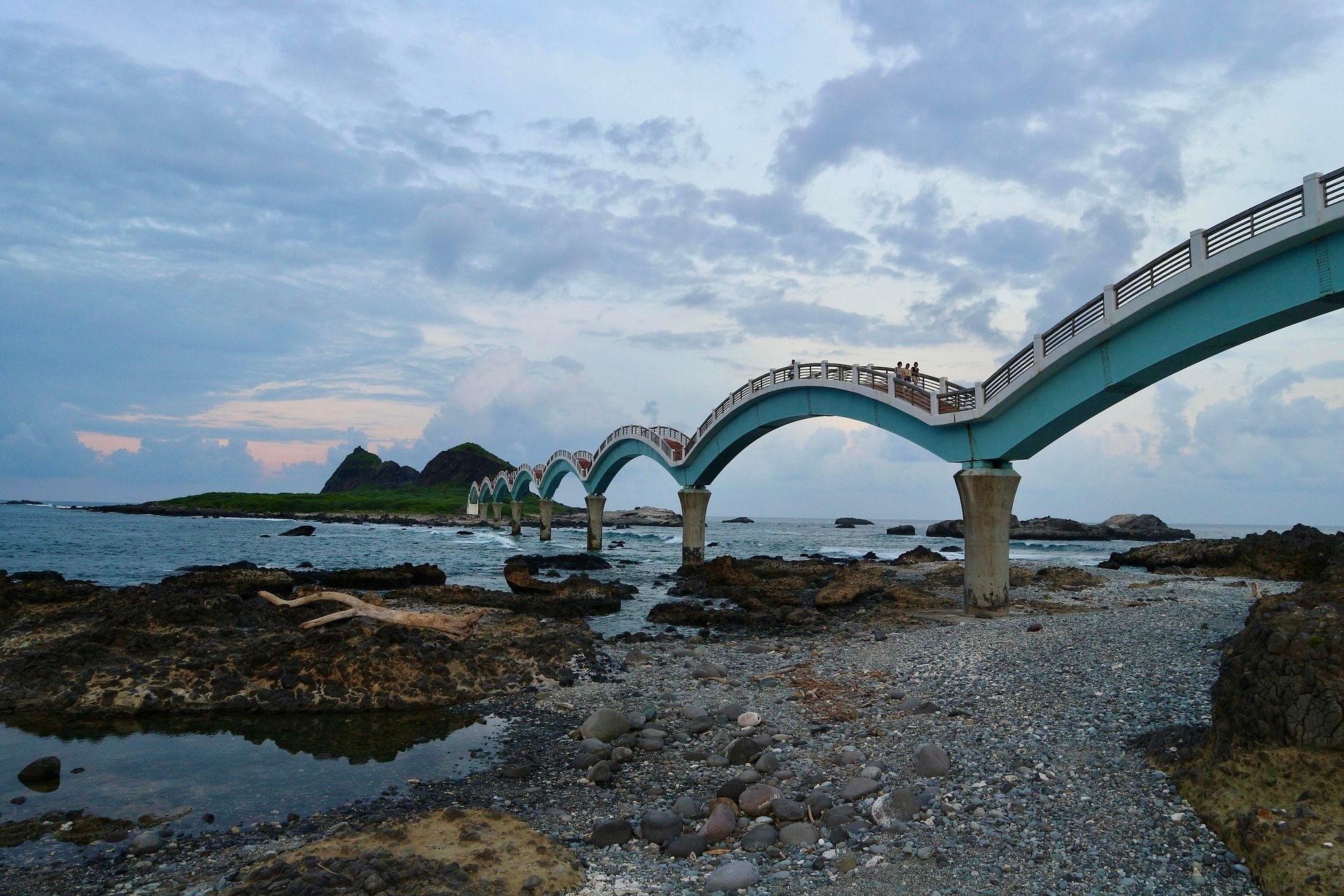 Eine hellblaue, mehrbogige Brücke erstreckt sich vom felsigen Ufer über das Meer zu einer kleinen Insel unter einem bewölkten Himmel.
