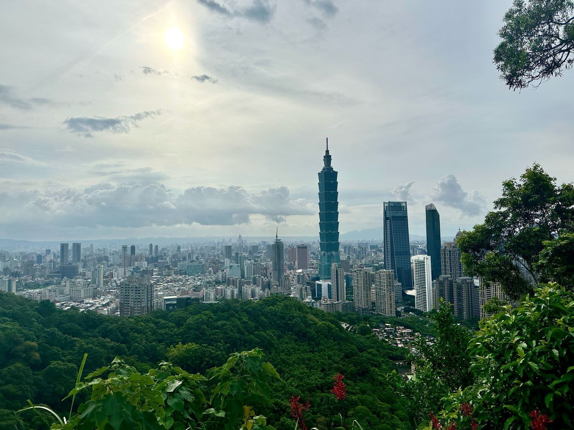 Eine Stadt-Skyline mit einem markanten hohen Wolkenkratzer, von oben über einem dichten grünen Wald unter einem hellen, bewölkten Himmel betrachtet.