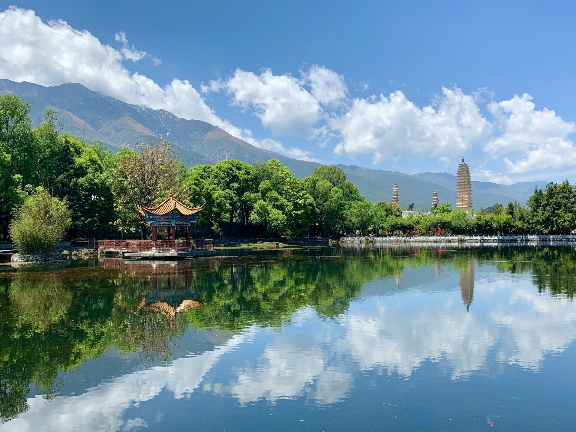 Un padiglione tradizionale e tre pagode si affacciano su un lago, riflettendo gli alberi verdi, le montagne distanti e il cielo azzurro e nuvoloso.