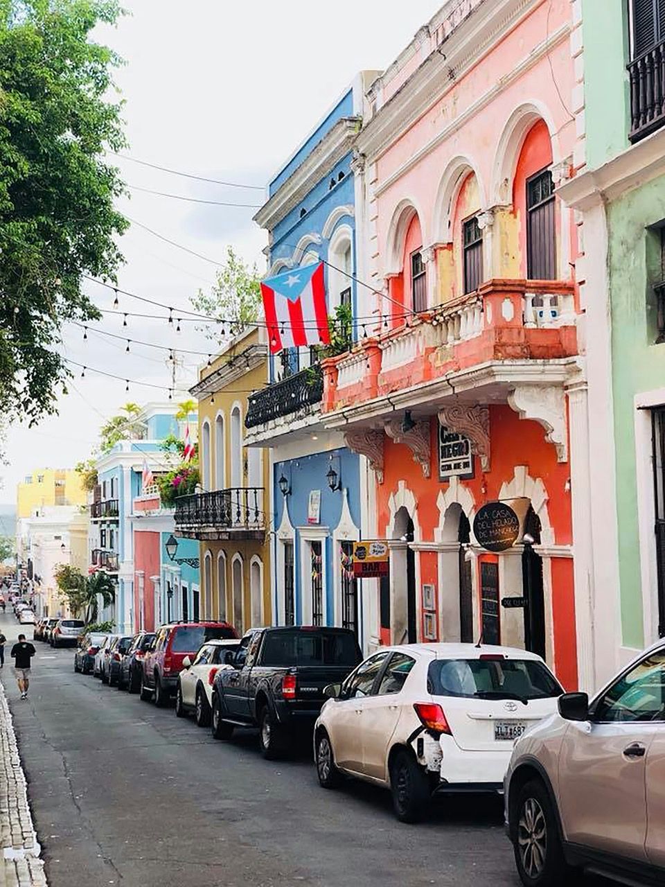 A Puerto Rican flag hangs from a balcony on a street lined with colorful buildings and parked cars.