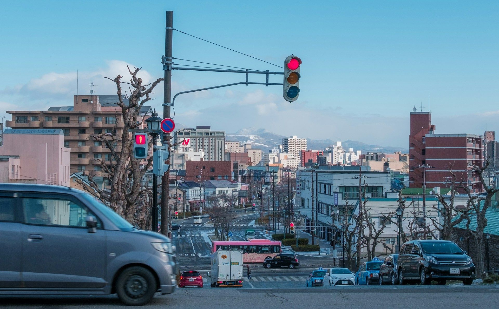 Una vista ampia di una strada cittadina giapponese con traffico, edifici e montagne innevate distanti sotto un cielo azzurro chiaro.