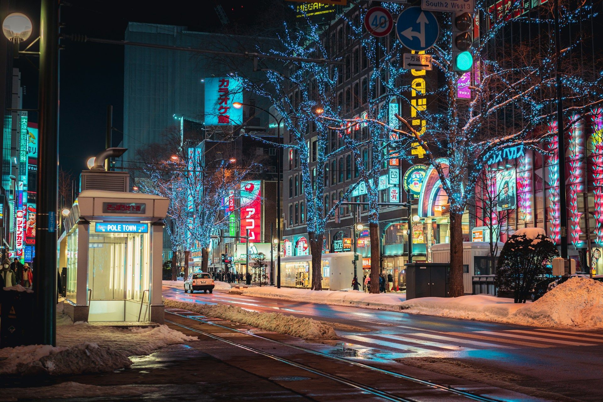 Una strada cittadina innevata di notte illuminata da insegne al neon colorate e alberi avvolti da luci blu, con binari del tram in primo piano.