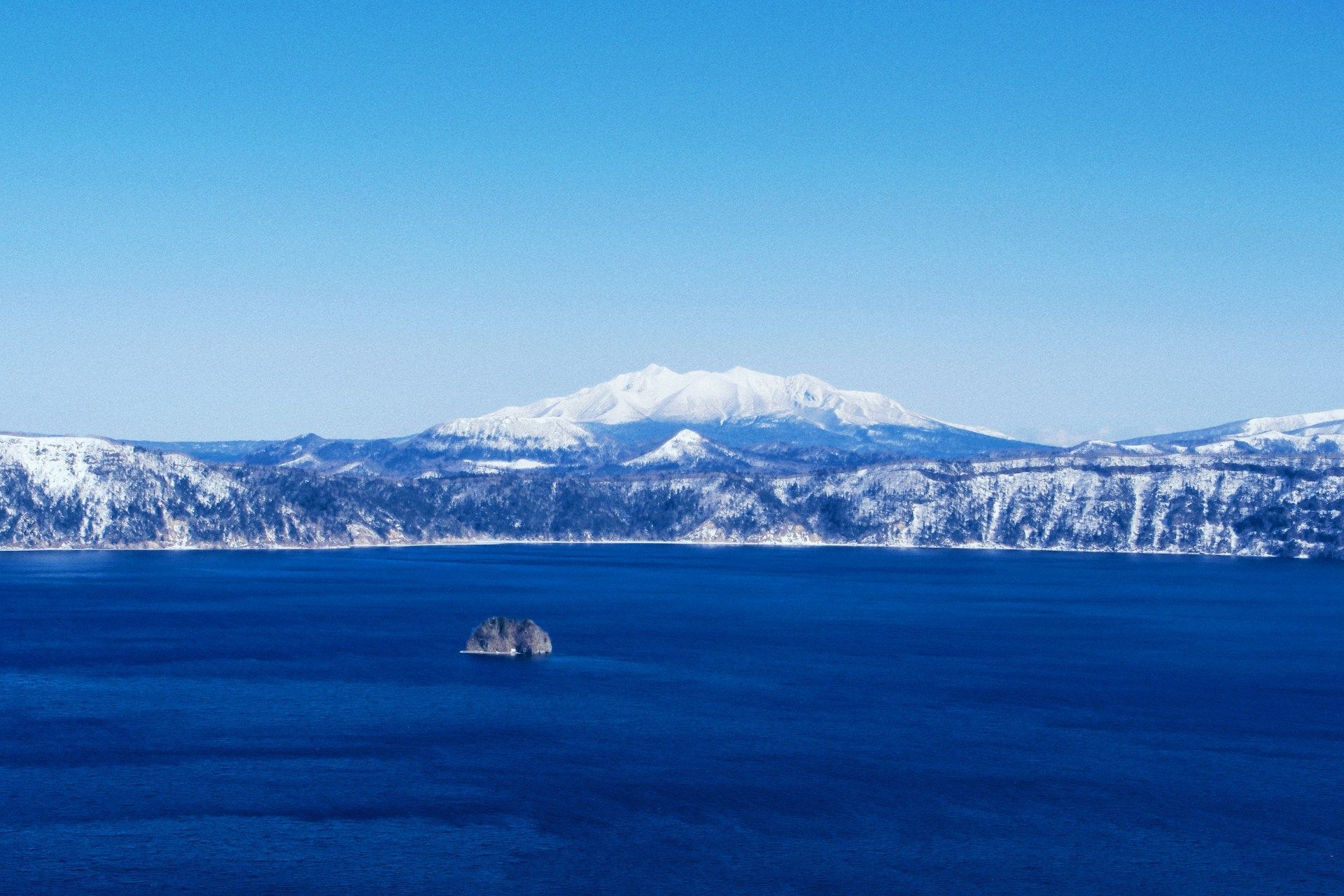 Un'ampia vista di un lago blu profondo con un piccolo isolotto roccioso, sullo sfondo montagne innevate sotto un cielo limpido.