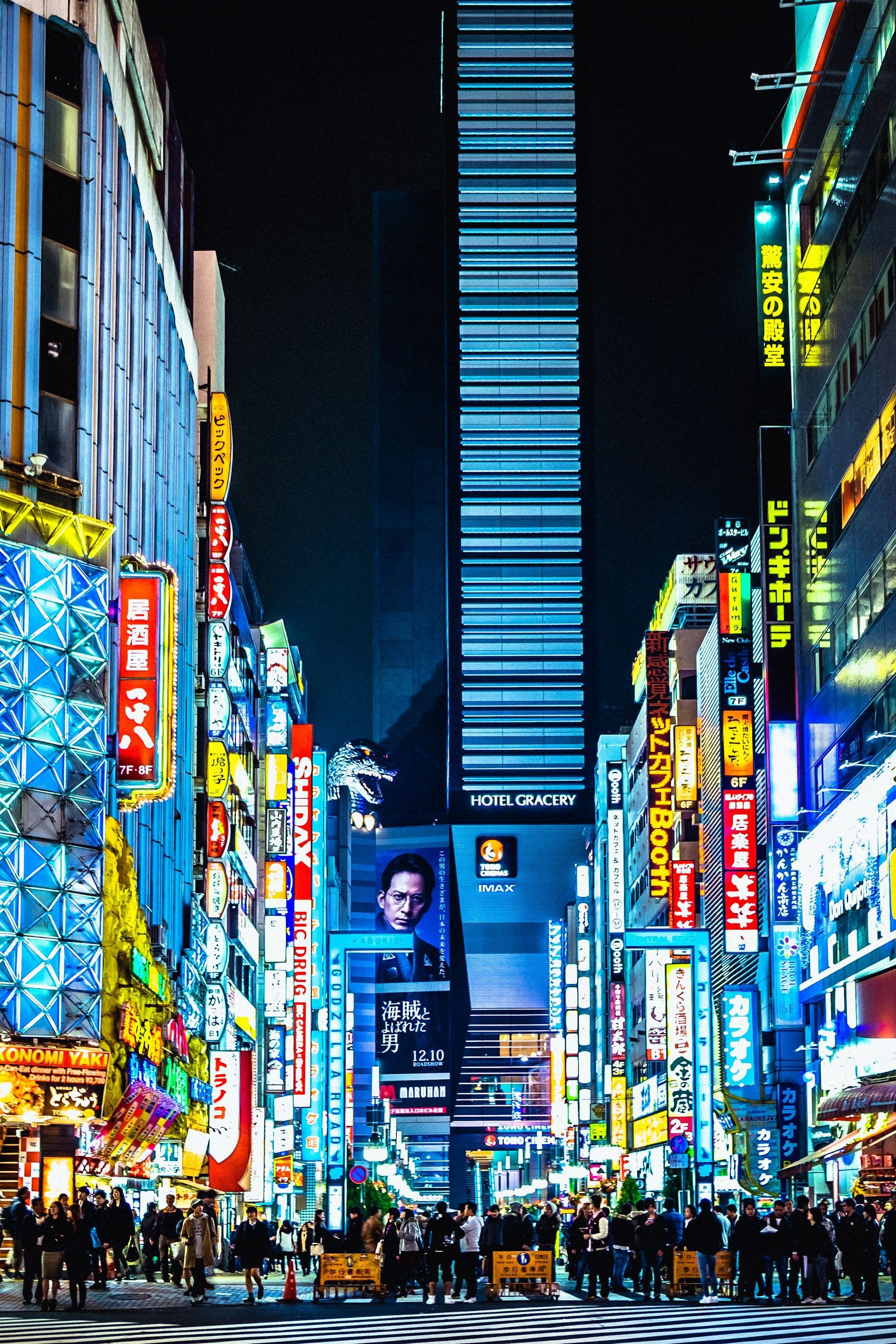 Una strada cittadina animata di notte, affollata di persone e illuminata da vivaci insegne al neon su edifici imponenti.
