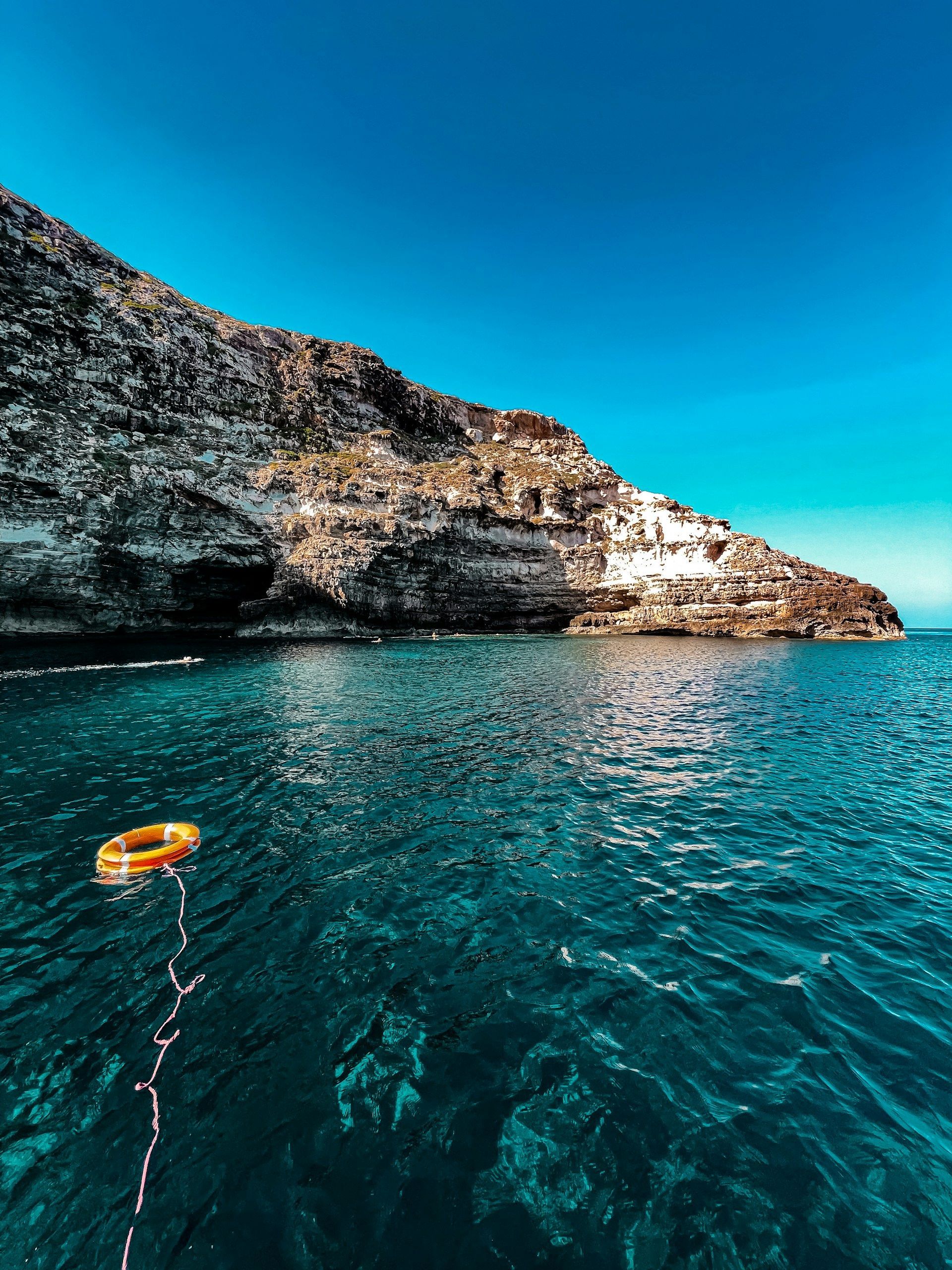 Un salvagente arancione galleggia sul mare turchese intenso accanto a una scogliera rocciosa sotto un cielo azzurro limpido.