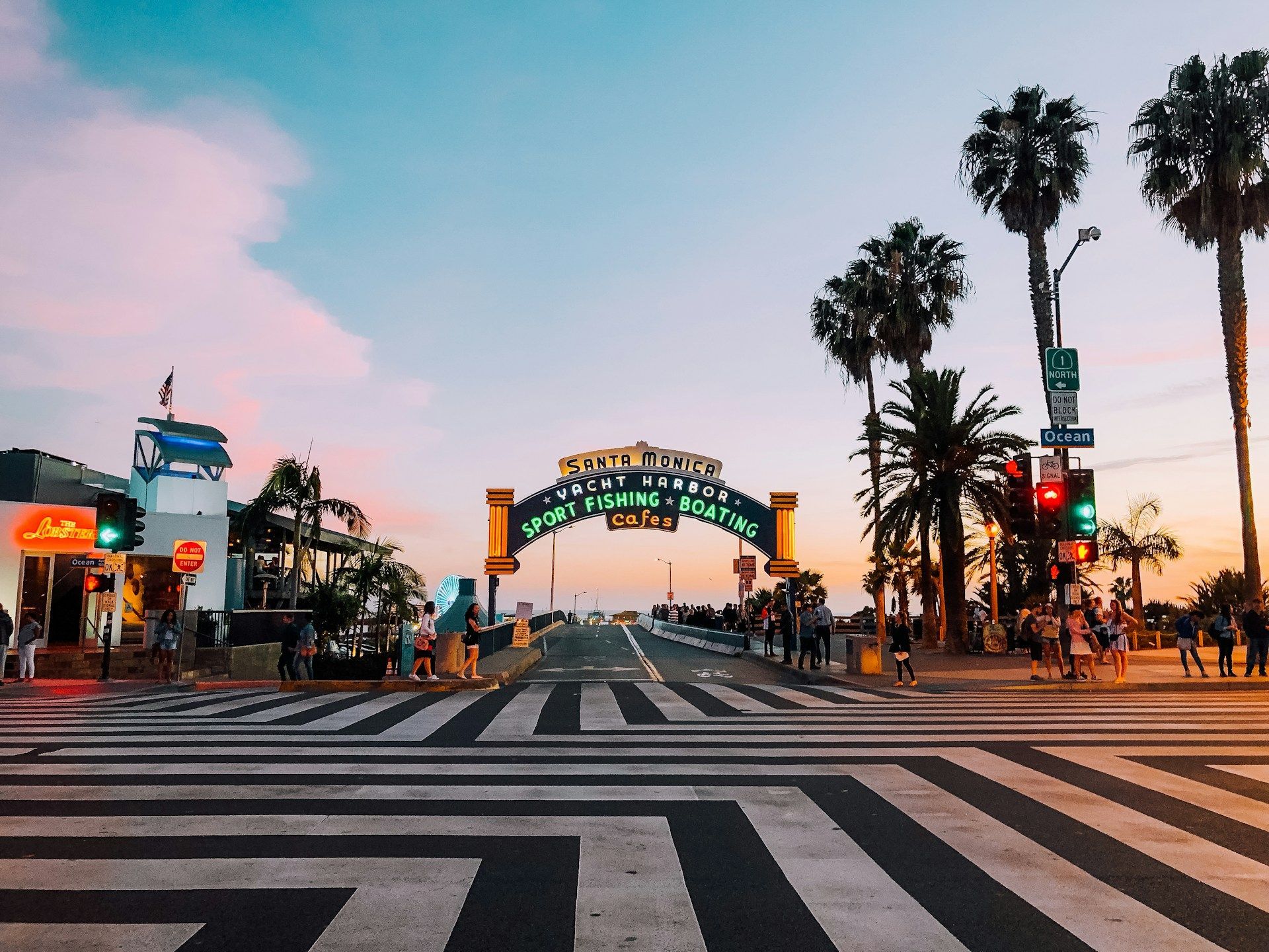 L'insegna luminosa dell'ingresso del molo di Santa Monica si erge ad arco sulla strada al tramonto, con palme e un attraversamento pedonale in primo piano.