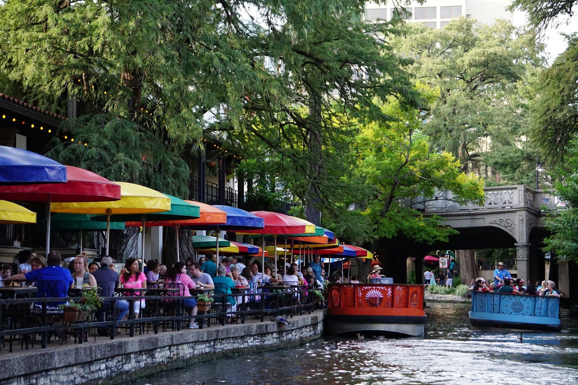 La gente mangia in un ristorante sul fiume sotto ombrelloni colorati mentre due battelli turistici navigano lungo il canale alberato.