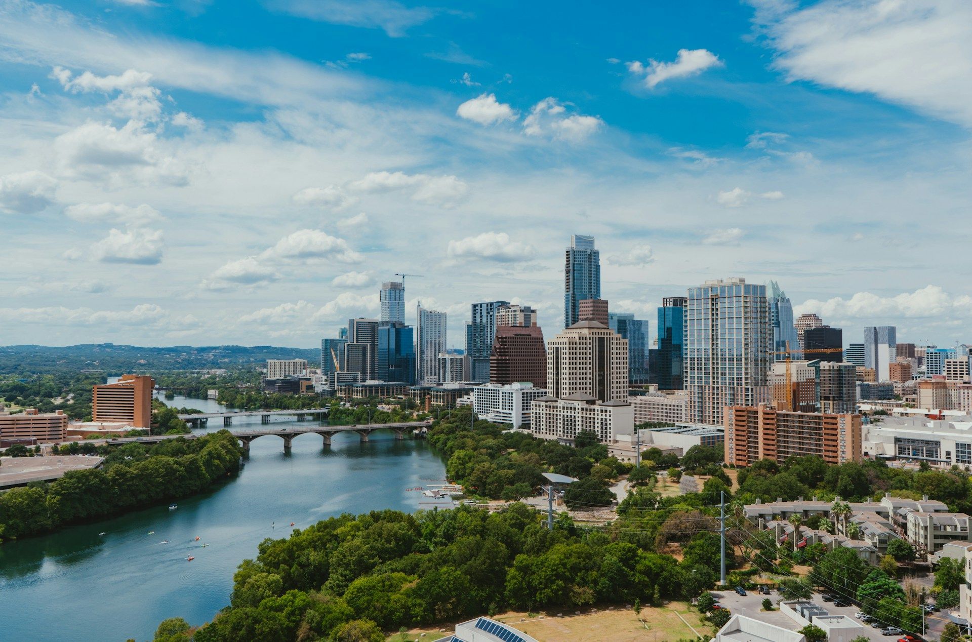 Uno skyline urbano con grattacieli si staglia dietro un ampio fiume, circondato da una vegetazione lussureggiante, sotto un cielo parzialmente nuvoloso.