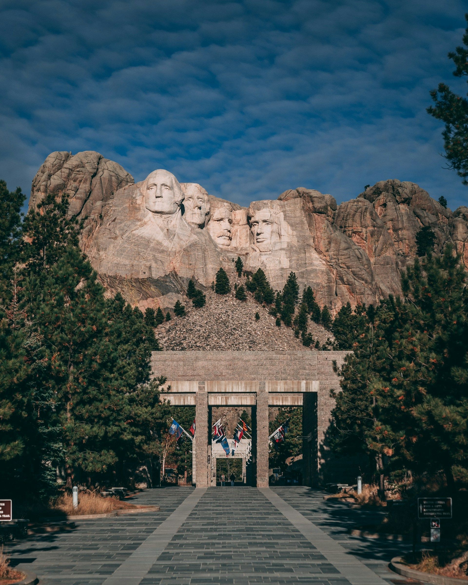 Il Monte Rushmore National Memorial visto dal vialetto d'ingresso in pietra, fiancheggiato da pini sotto un cielo azzurro.