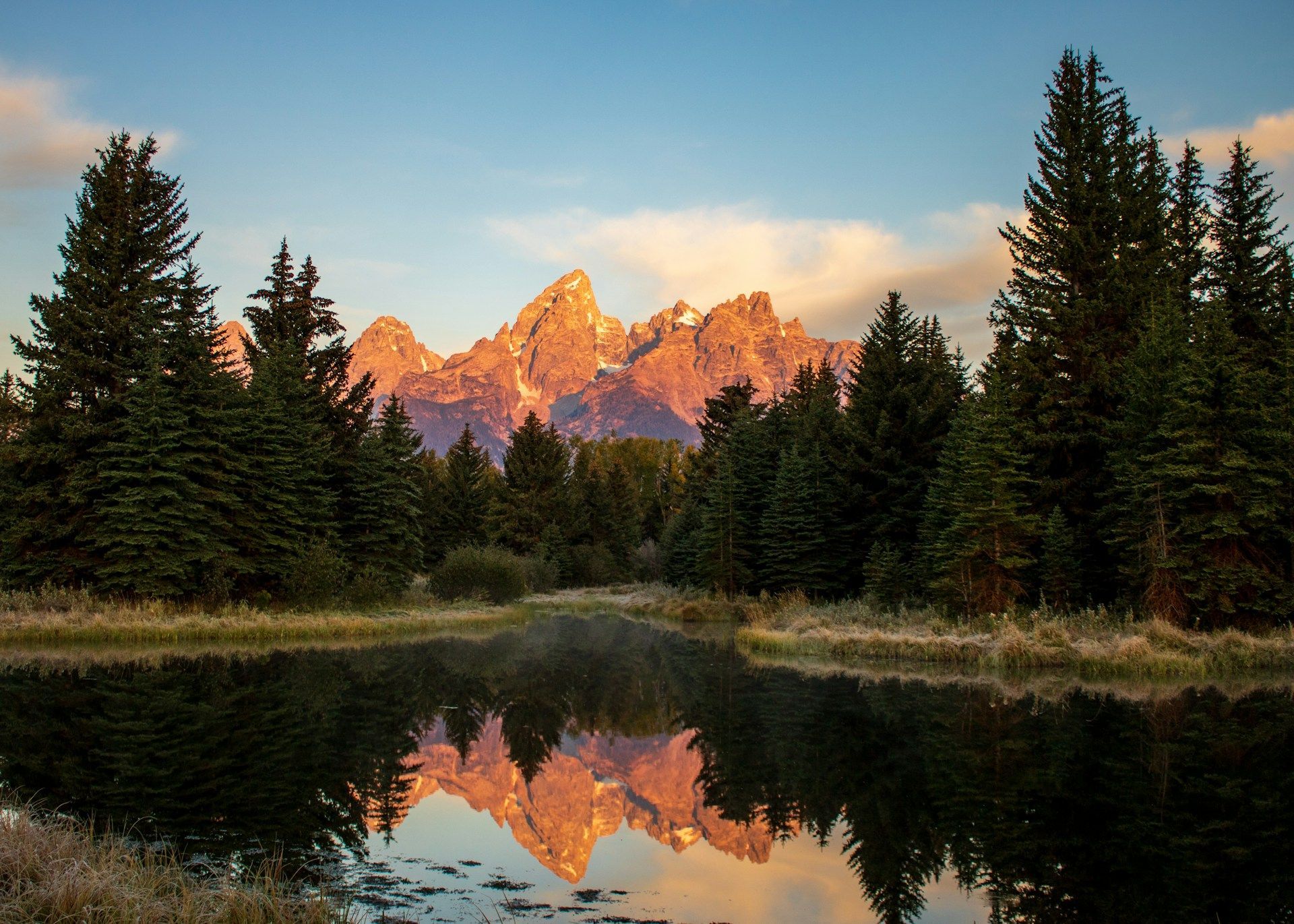 Cime montuose frastagliate illuminate dal sole si riflettono in un lago immobile, con una fitta foresta sempreverde in primo piano.