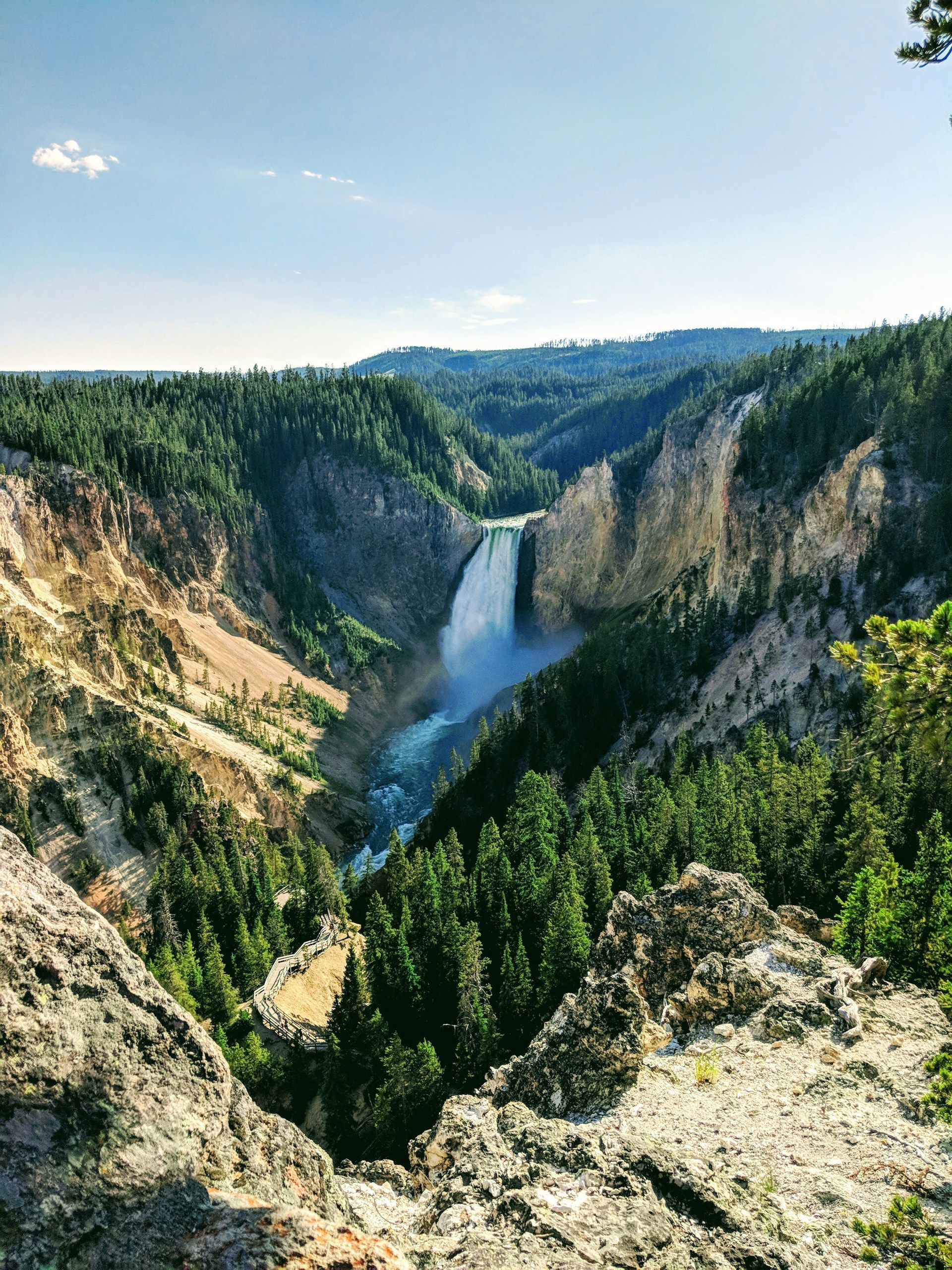 Una grande cascata precipita in un fiume all'interno di un vasto canyon densamente boscoso di pini sotto un cielo azzurro.