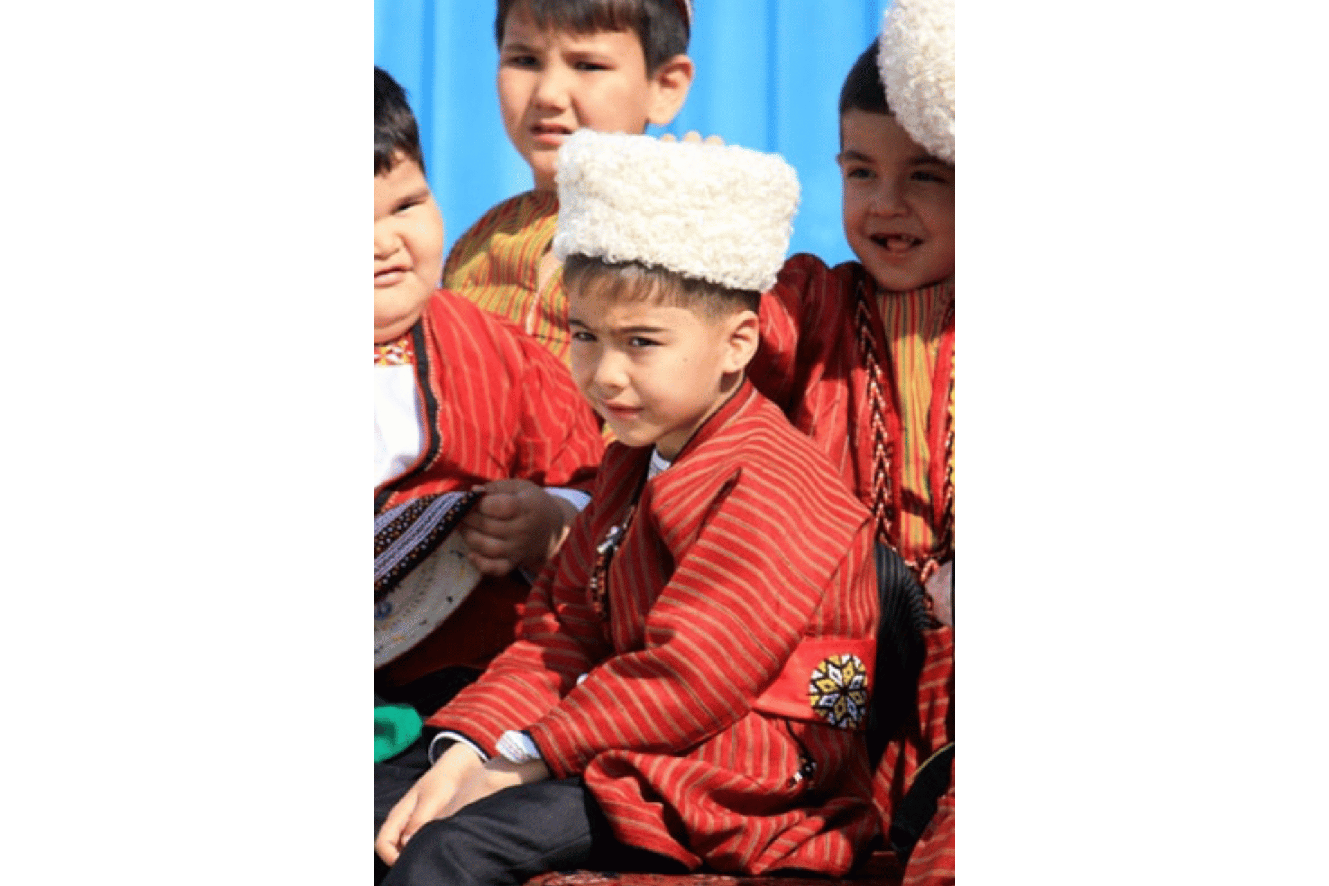 Un bambino, con una giacca tradizionale a righe rosse e un cappello di lana bianco, siede in un gruppo di bambini, guardando la fotocamera.