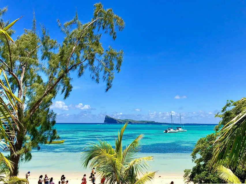 Una vista da una spiaggia di sabbia, un oceano turchese con un catamarano e un'isola rocciosa all'orizzonte, incorniciata da palme.